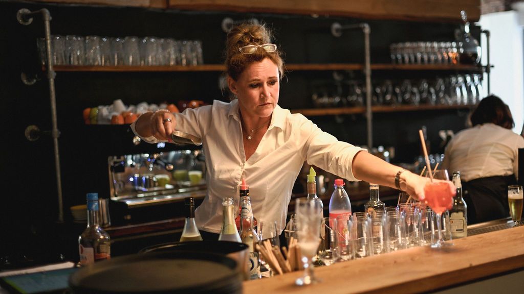 A bartender pours drinks at a modern bar counter with shelves of glassware and bottles in the background.