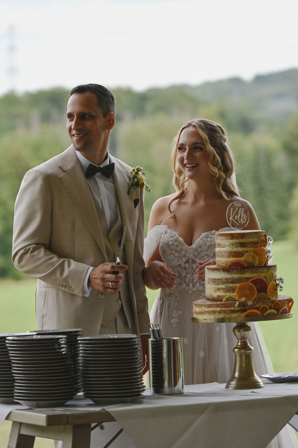 A couple shares a romantic moment while cutting their rustic naked wedding cake at an outdoor reception.