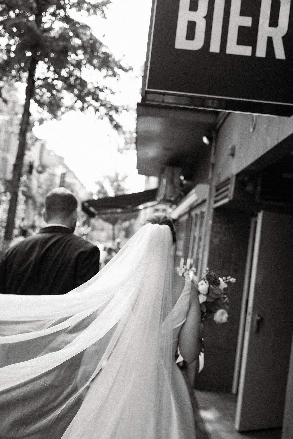 A flowing white wedding veil billows in the breeze as a couple walks past an urban storefront with black signage.