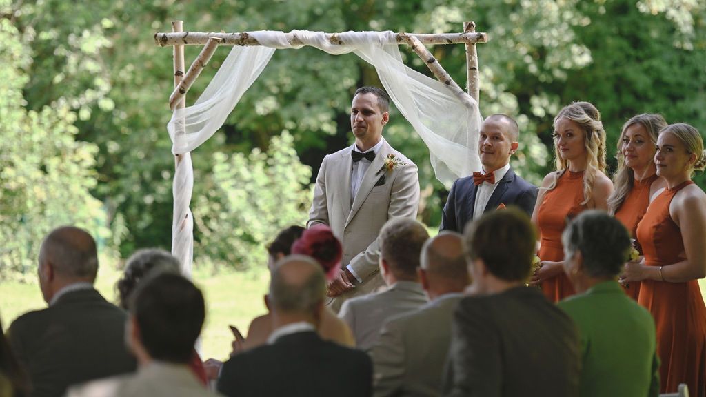 Wedding ceremony taking place outdoors under a wooden arch decorated with white fabric during a sunny summer day.