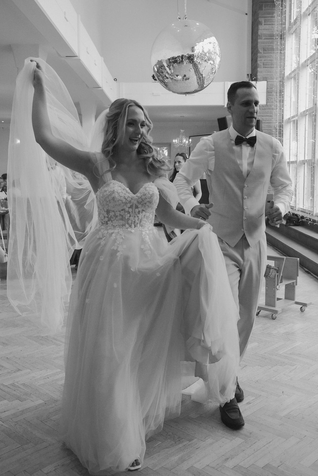 A joyful wedding party dances together in a bright indoor venue with hardwood floors and tall windows.