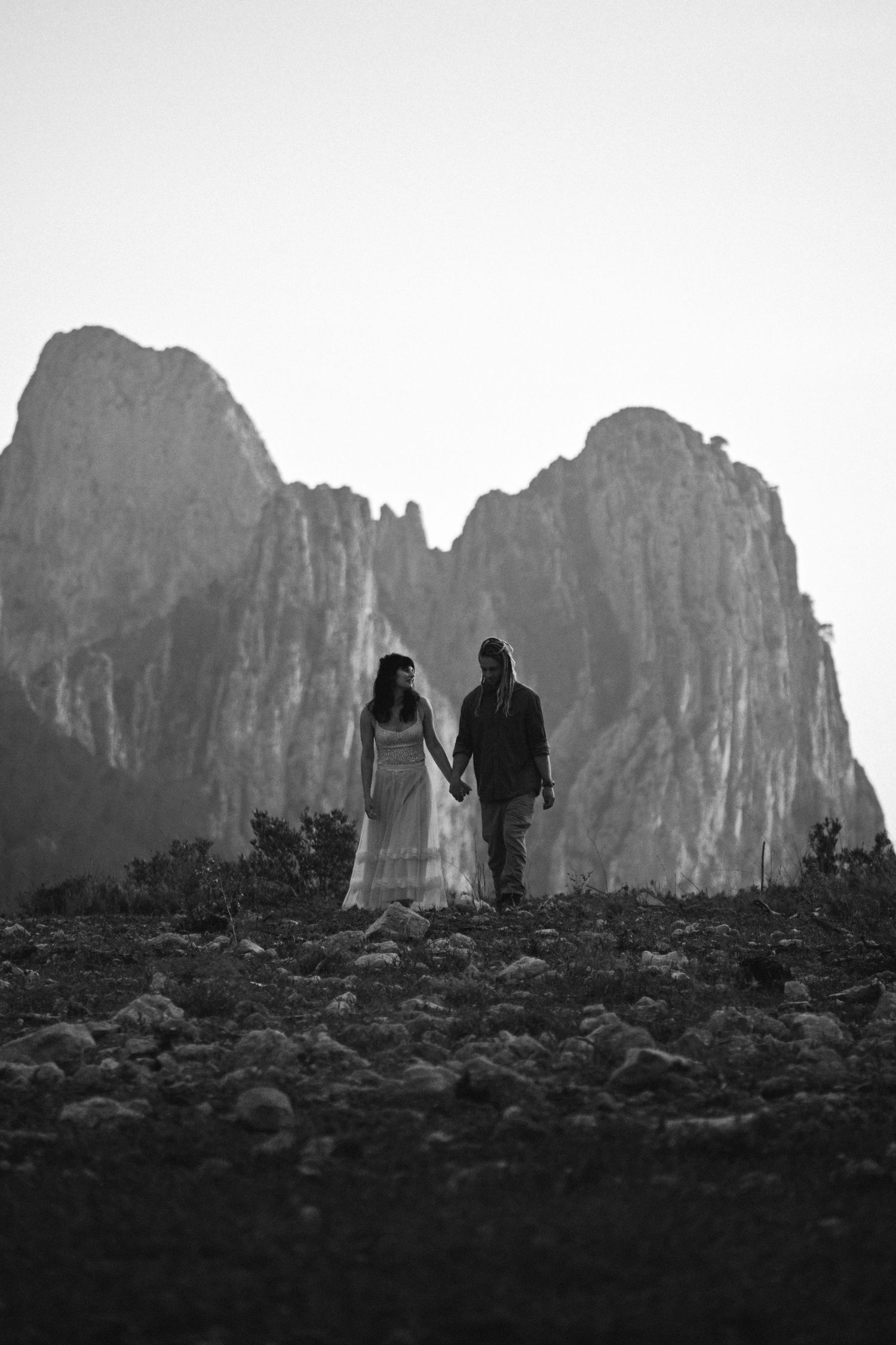 Black and white silhouette of couple holding hands against dramatic mountain backdrop.