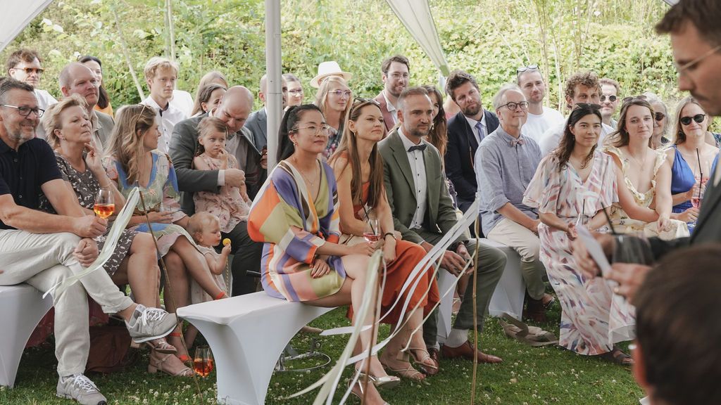Wedding guests seated in rows on white chairs under a tent canopy during an outdoor celebration.