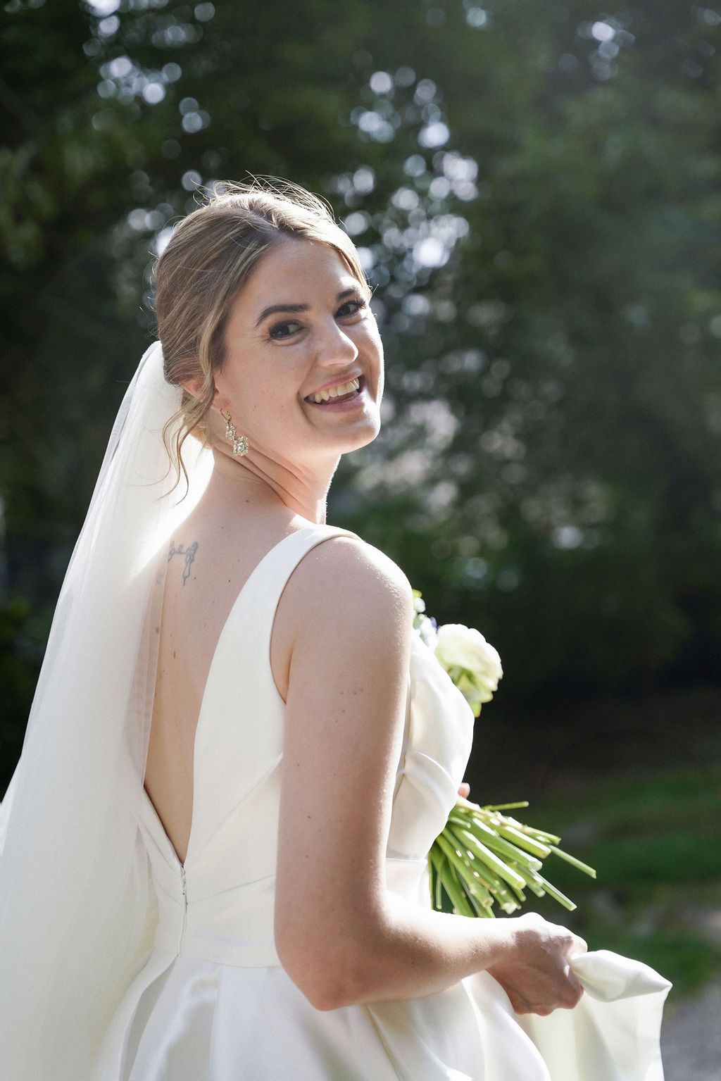 A joyful bride in a white wedding dress laughs while holding a bouquet outdoors.