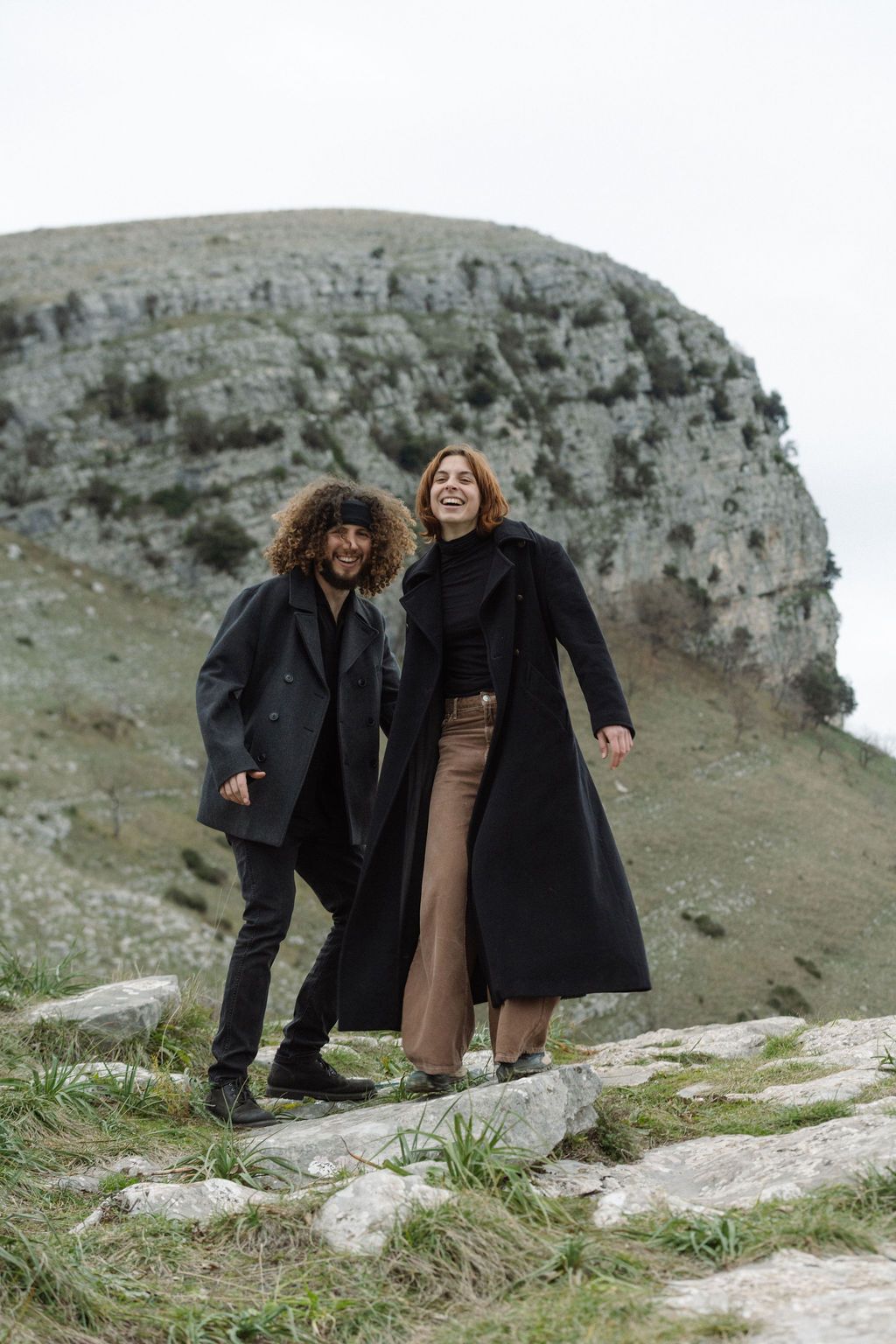 Two friends in long black coats standing on rocky terrain with cliffs in the background.