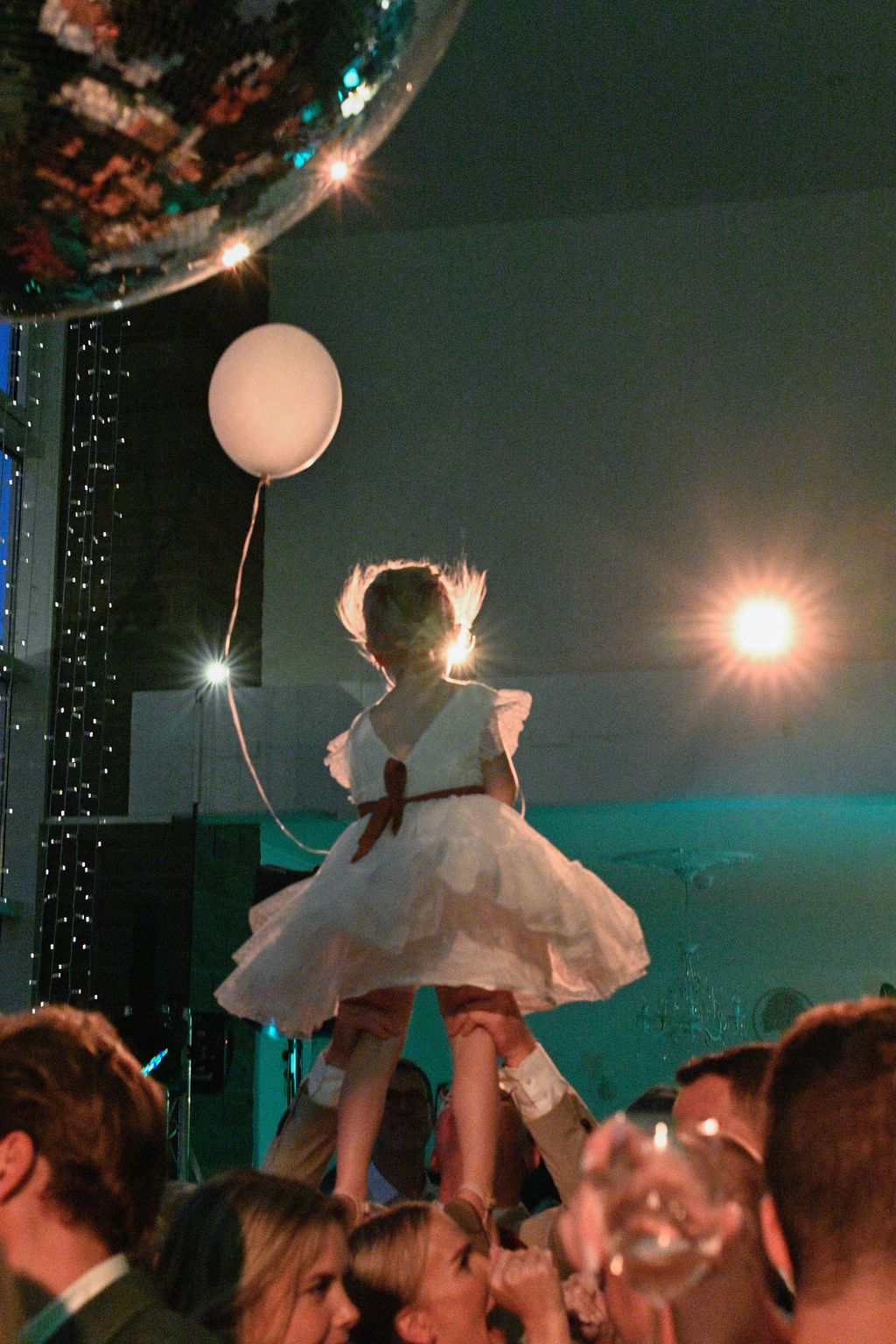 Wedding celebration with crowd lifting someone in white dress during reception party with balloons and twinkling lights.