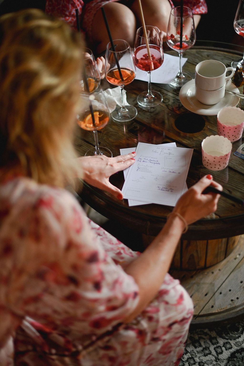 Person in floral dress writes notes while enjoying wine at rustic wooden table with glassware and papers.