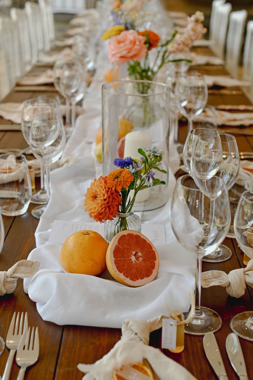 Elegant table setting with citrus fruits and orange flowers decorating a wooden dining table with white linens and wine glasses.