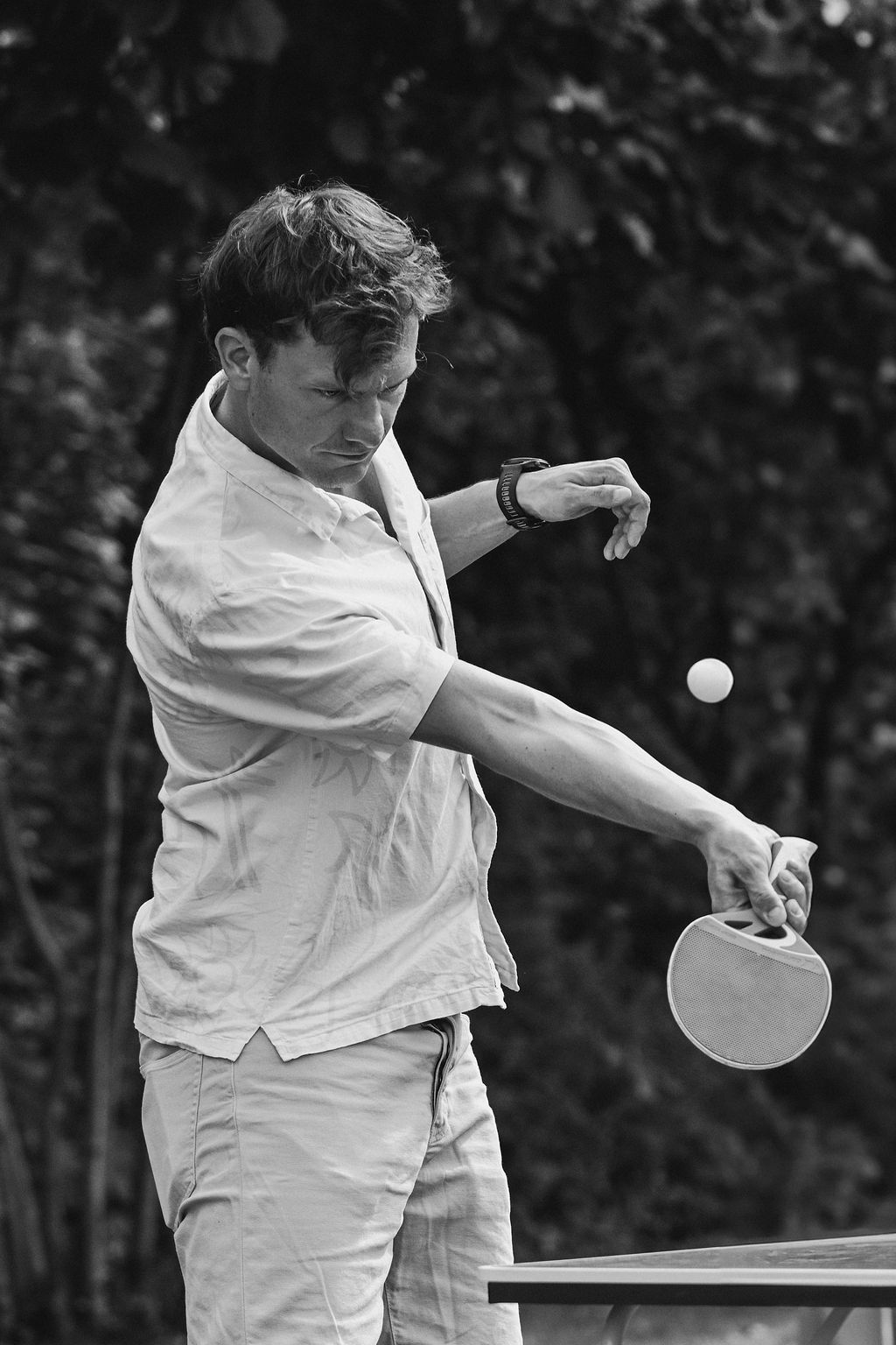 Person in white clothing playing table tennis with focused concentration at an outdoor venue.