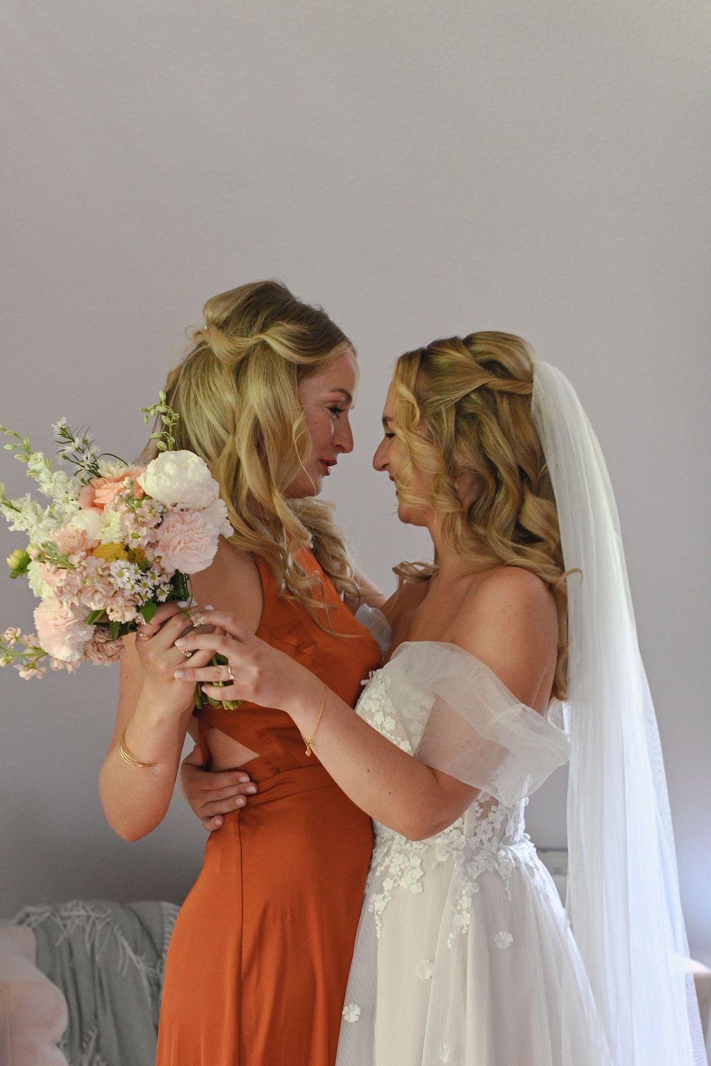 Two women in orange and white dresses share a playful moment with floral bouquets during a wedding photoshoot.