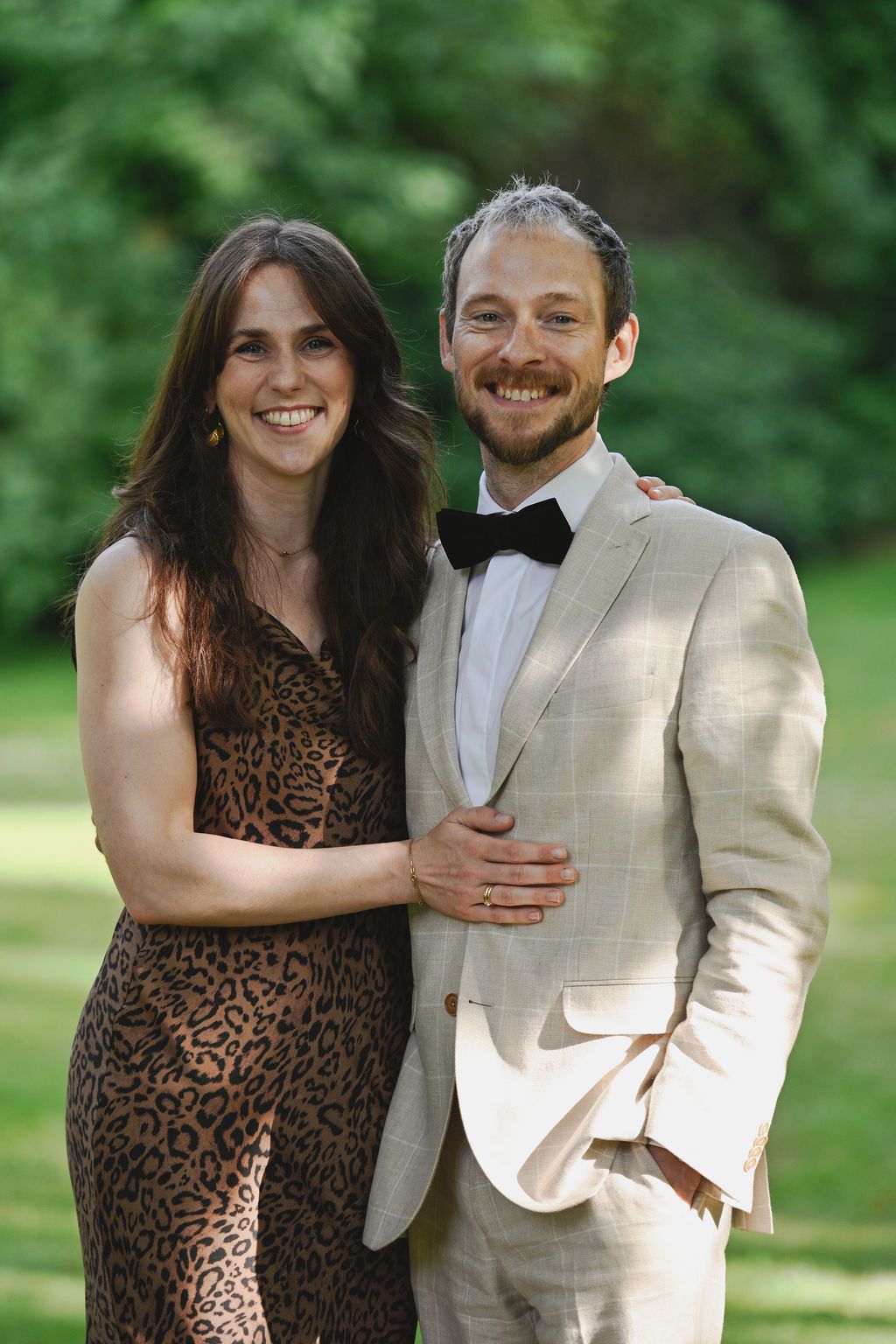 A couple poses outdoors in a garden setting wearing a leopard print dress and beige suit.