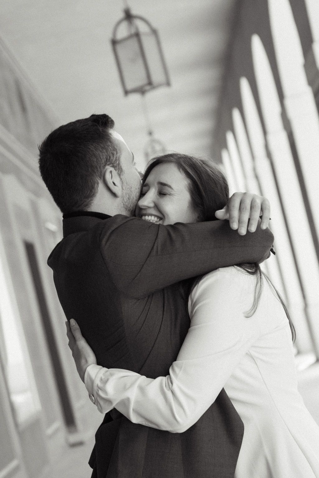 Black and white photo of a joyful couple embracing and smiling in an elegant columned hallway.