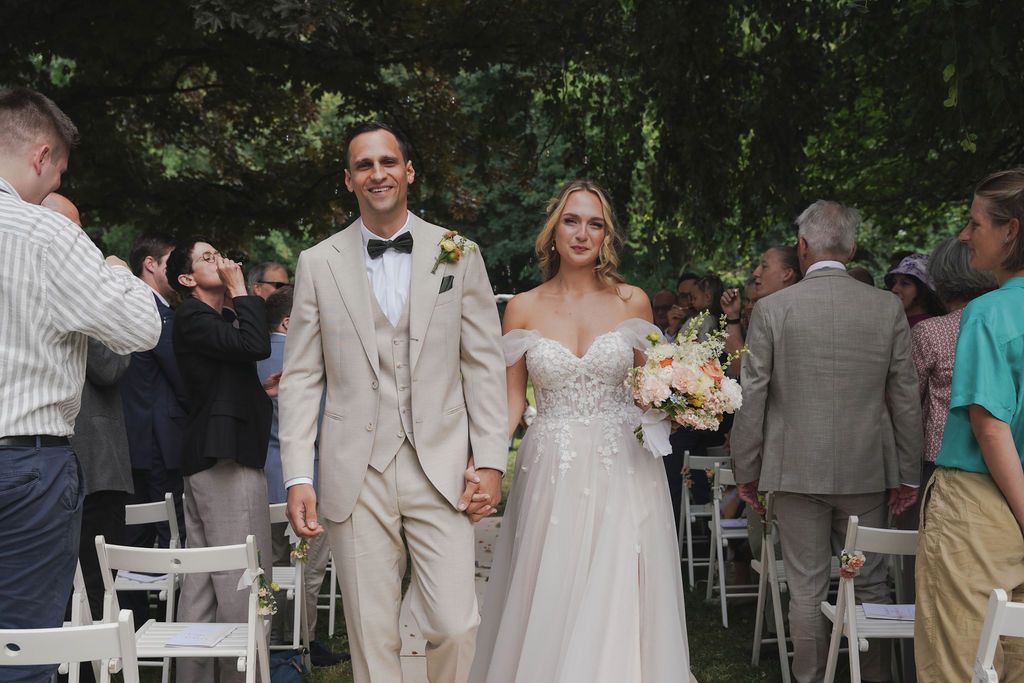 Wedding guests gather outdoors for a celebration under trees with white chairs and decorations.