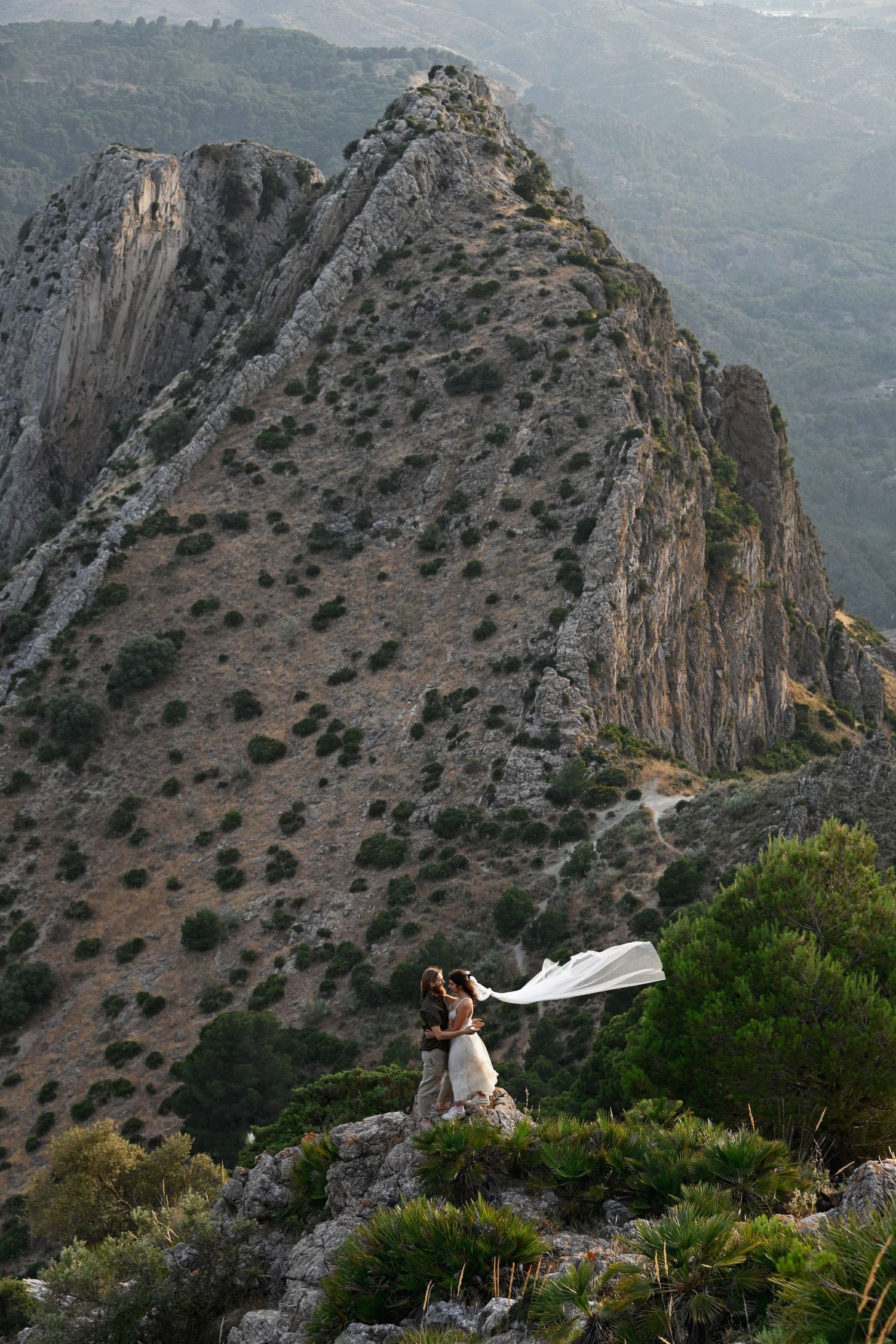 Wedding couple stands on dramatic mountain ridge with flowing white dress in wind.