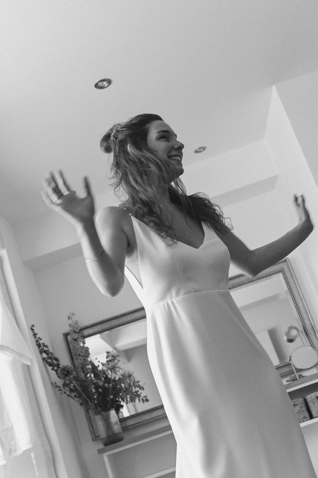 Black and white photo of someone dancing joyfully in a white dress in a bright interior room with a mirror.