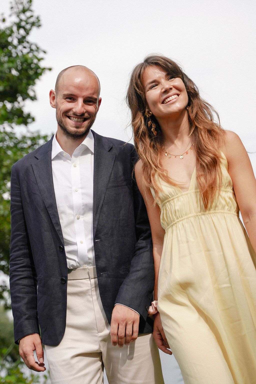 A couple in casual wedding attire share joyful candid moments outdoors, with her in a yellow sundress and him in a blazer.
