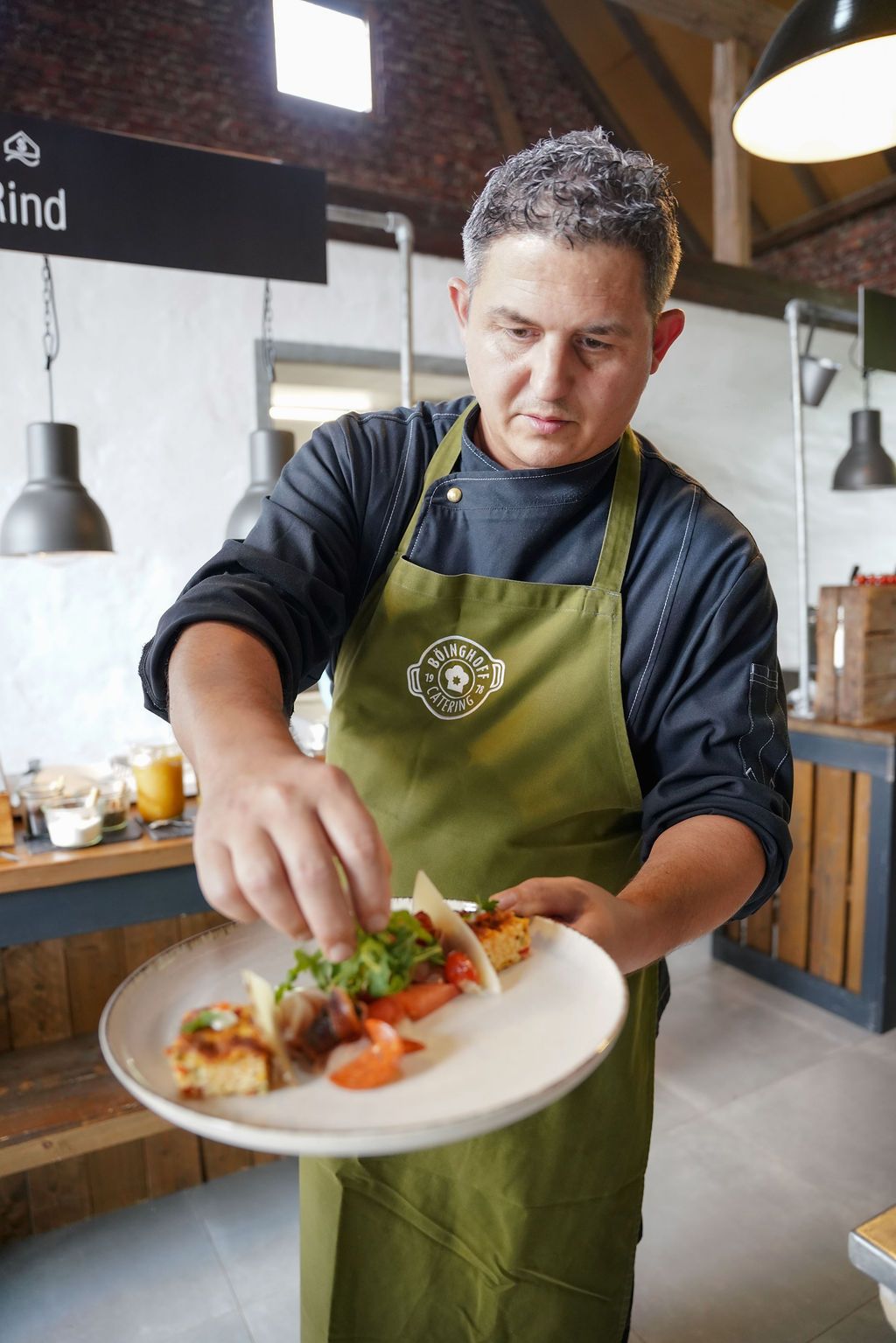 Chef in green apron plates gourmet dishes in restaurant kitchen with brick walls and pendant lighting.