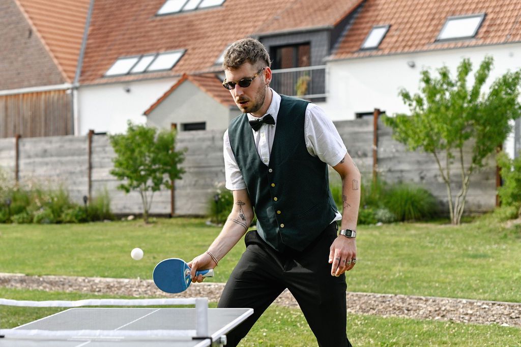 A person in formal attire plays ping pong on an outdoor table near a residential building.