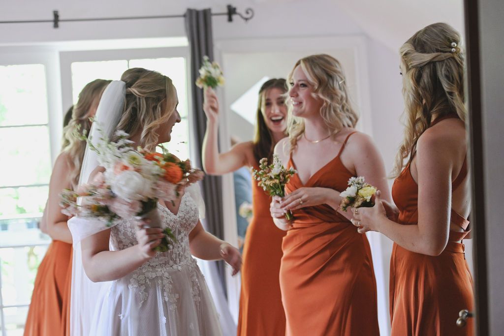 Bride in white dress shares joyful moment with bridesmaids wearing burnt orange dresses during wedding preparations.