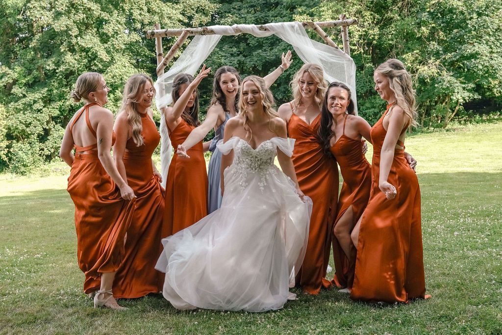 Wedding party in burnt orange dresses poses playfully on green lawn during outdoor celebration.
