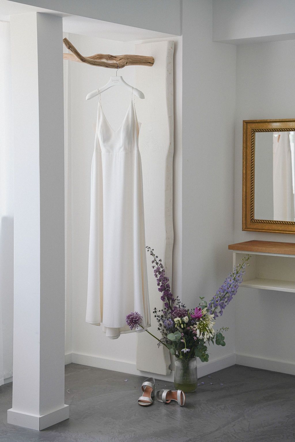 A white wedding dress hangs in a minimalist dressing room with a floral arrangement and sandals on the floor.
