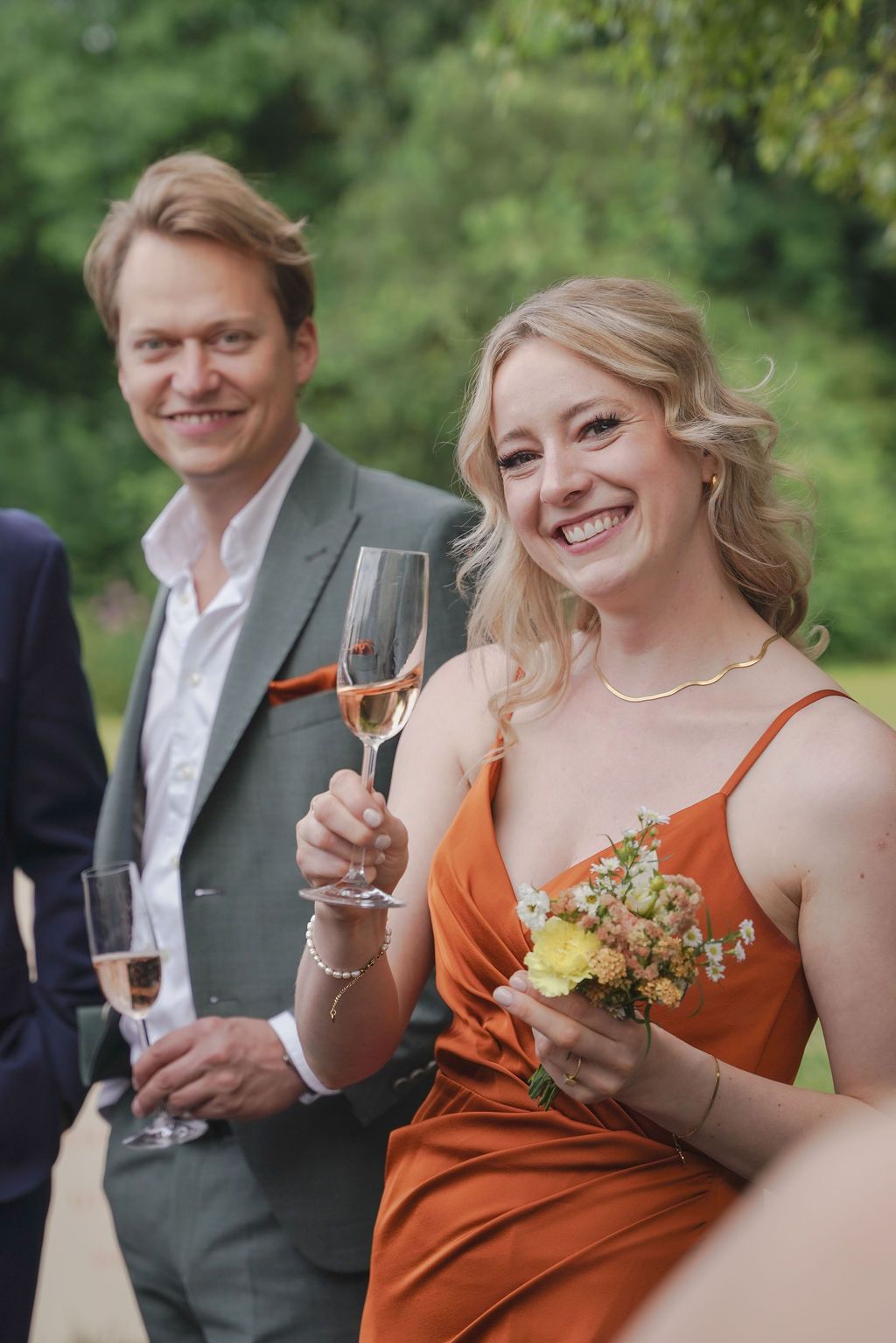 A couple in wedding attire share a toast outdoors, with the person in orange holding a champagne glass.