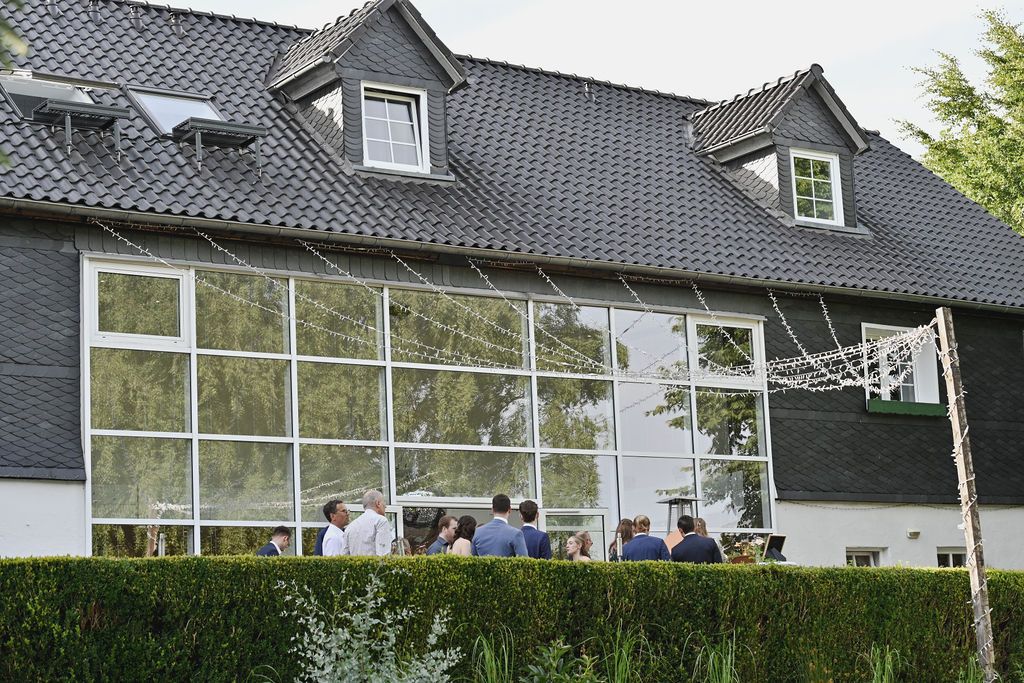 Modern dark gray house with large glass windows and people seated on a patio area outside.