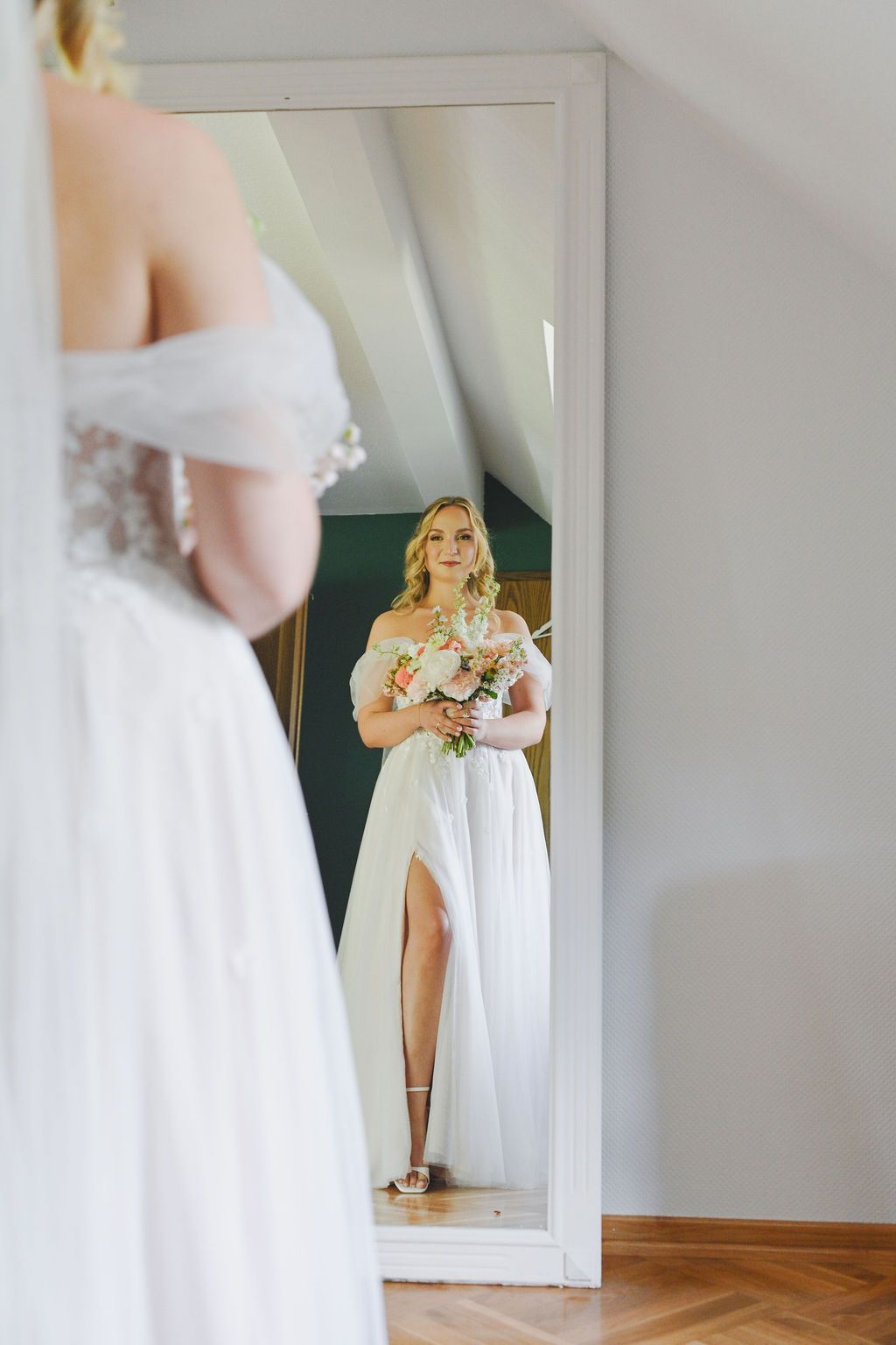 A bride in an elegant white off-shoulder wedding dress with a thigh slit looks at her reflection in a full-length mirror.