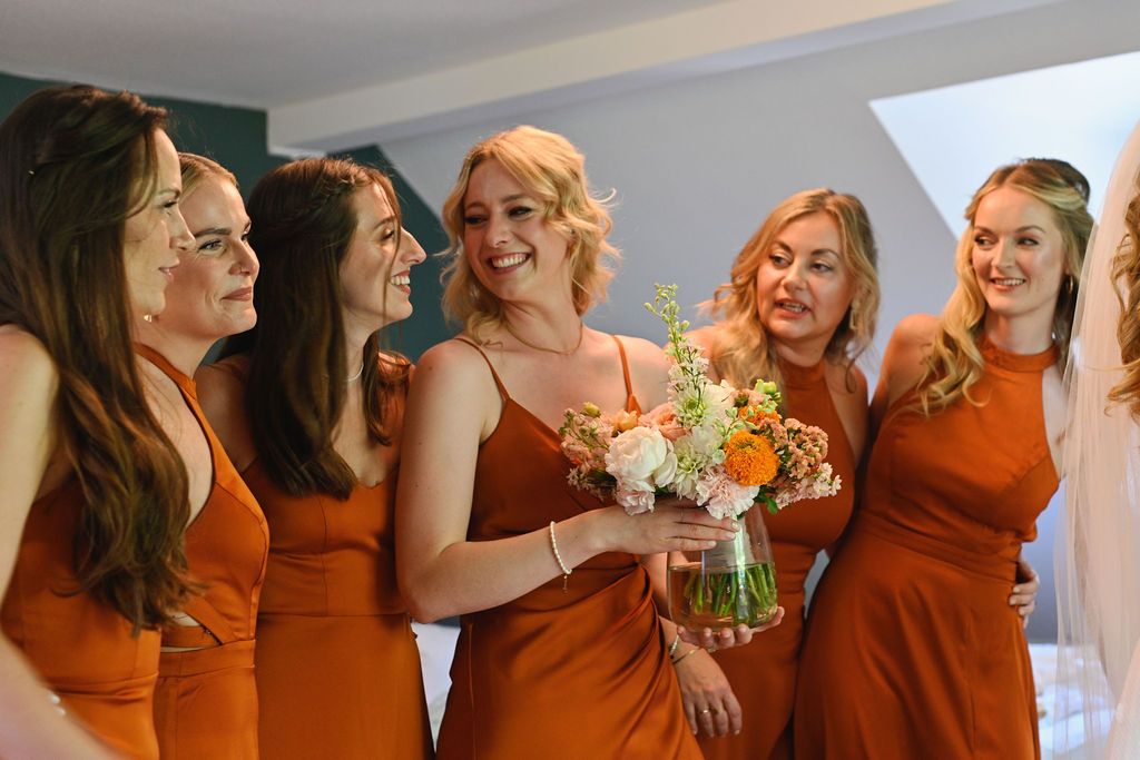 Bridesmaids in matching burnt orange dresses share laughter and hugs during wedding preparations.