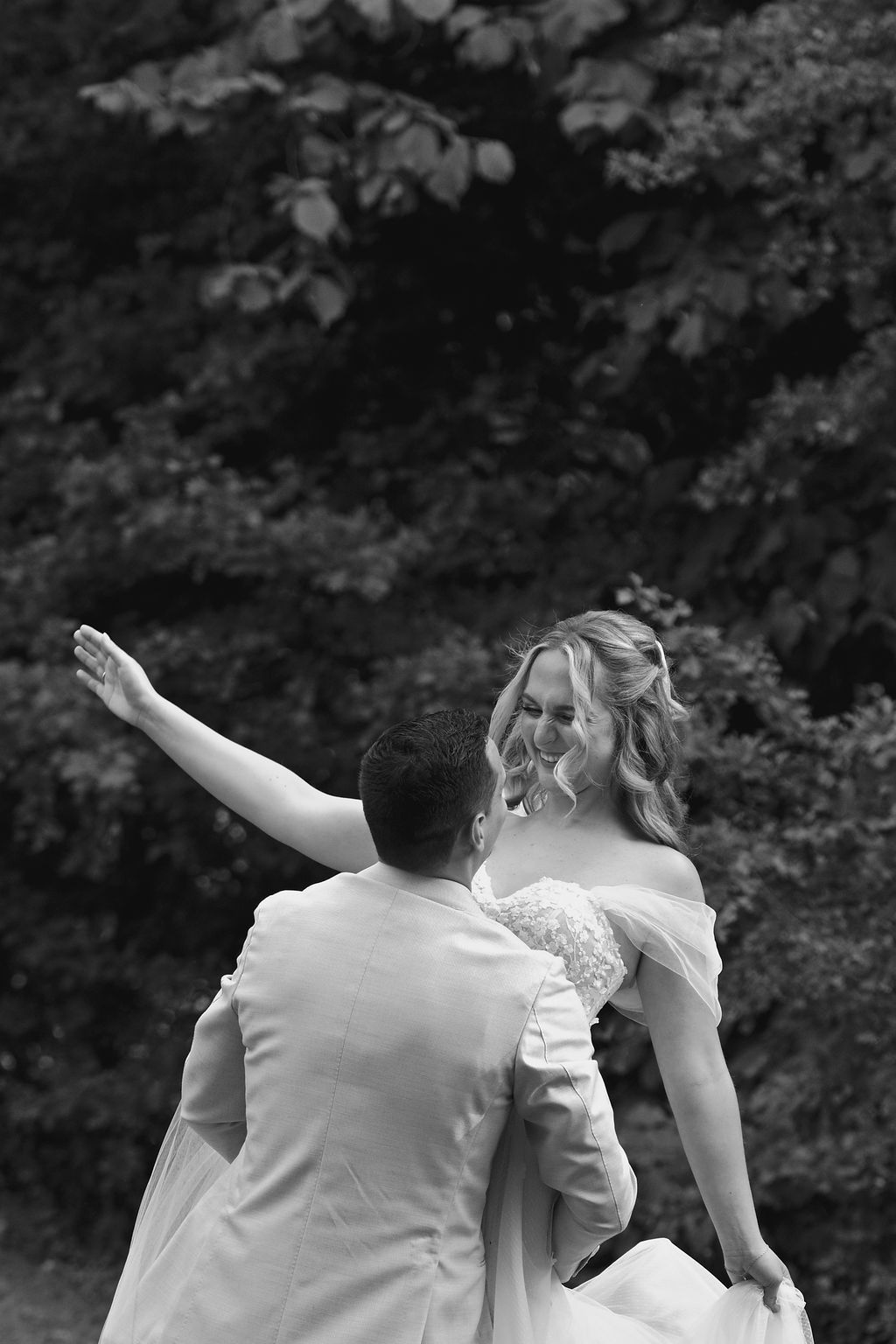 A couple in white wedding attire poses with their backs to the camera while making celebratory gestures in a garden setting.