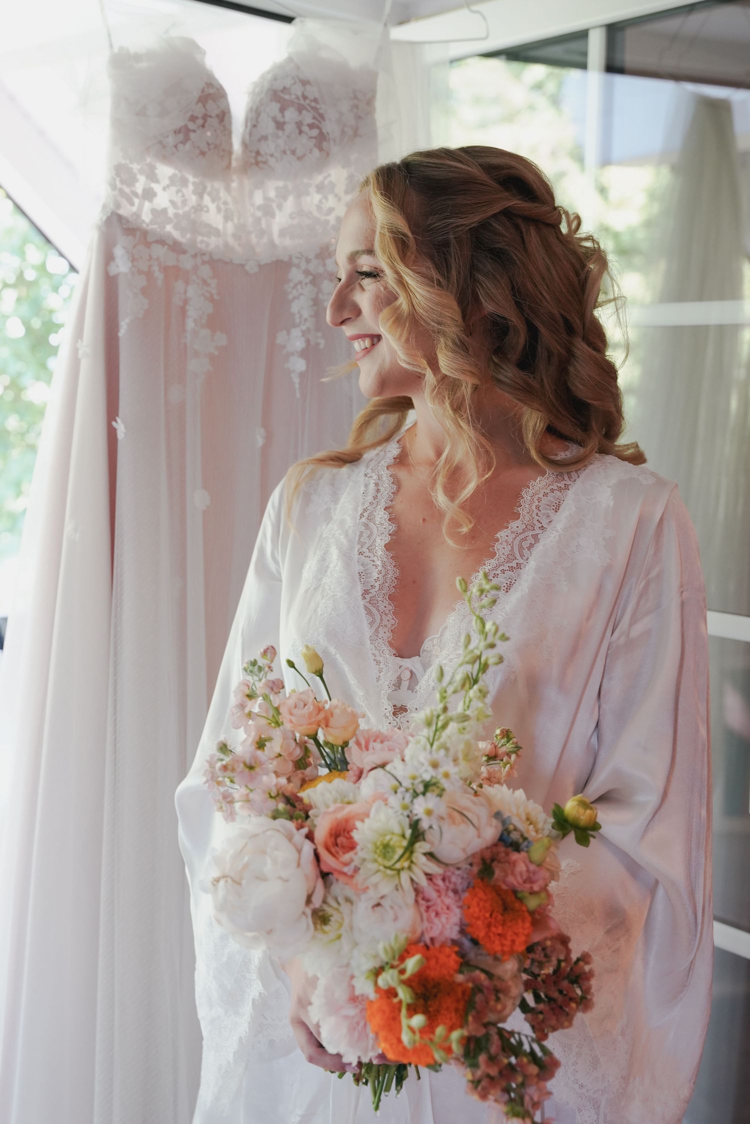 A bride in a white lace robe holds a vibrant bouquet of coral and peach flowers while her wedding dress hangs behind her.