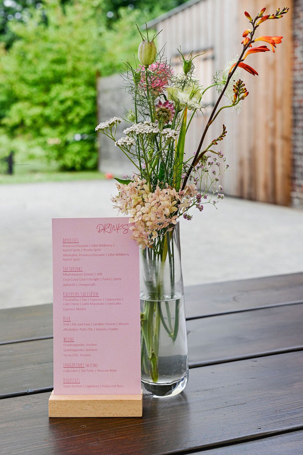 Small floral arrangement in glass vase next to pink menu card on dark wooden table with outdoor setting.