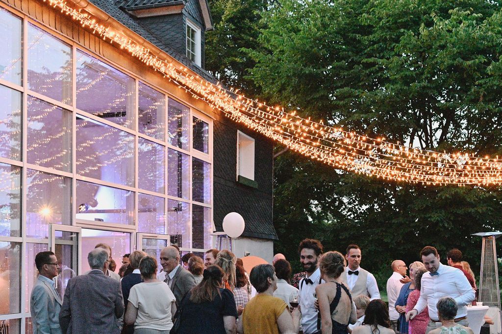 Wedding guests mingle at an outdoor evening reception with string lights illuminating a glass-walled venue.