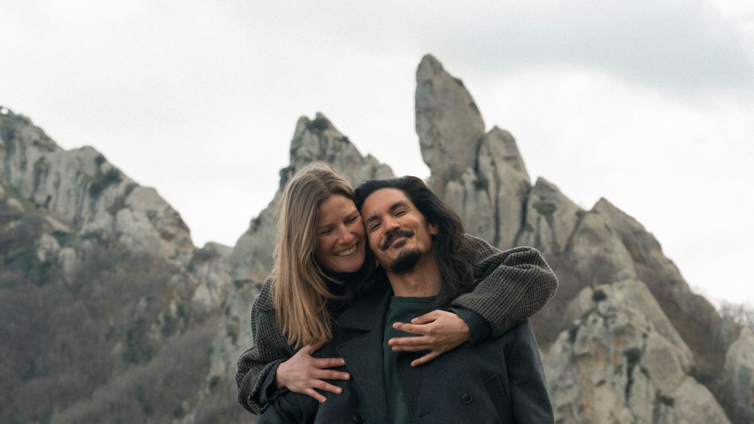 A happy couple embracing and smiling in front of dramatic rocky mountain peaks under a cloudy sky.