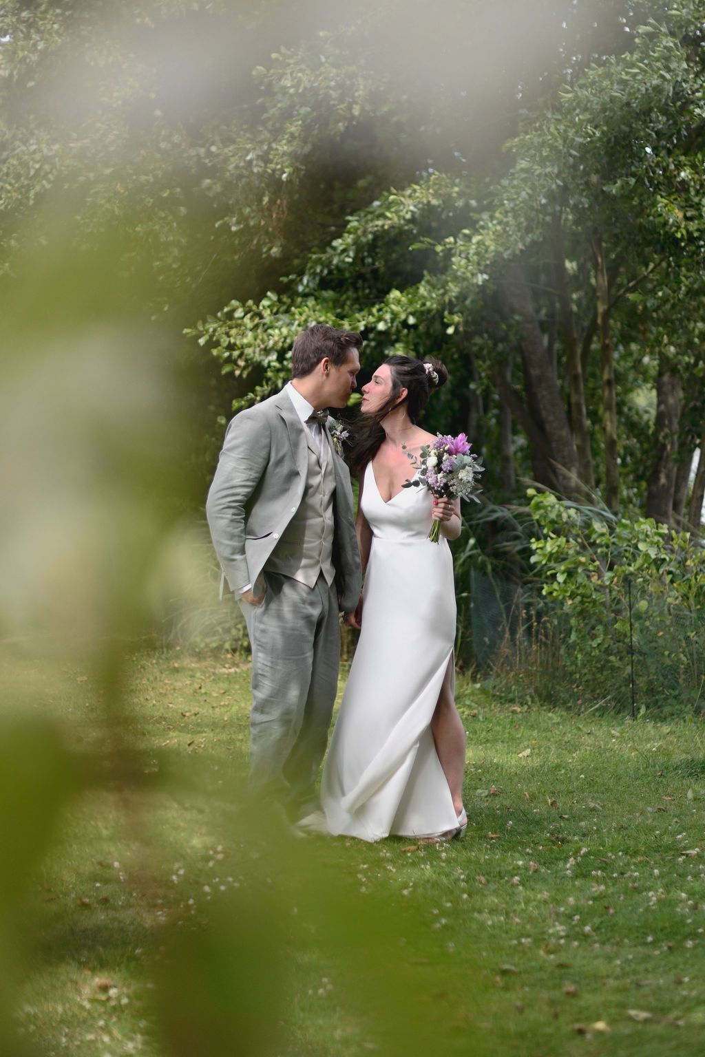 A bride and groom share a romantic moment in a lush garden, she holds a purple bouquet wearing a white gown, he wears a grey suit.
