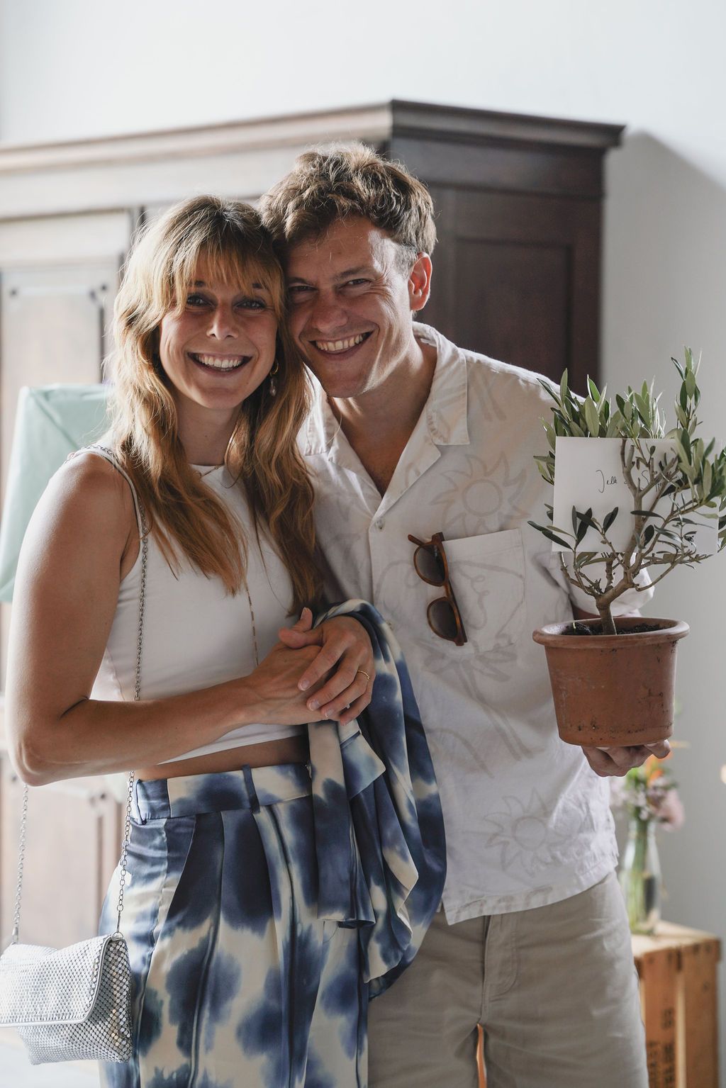A couple in white summer outfits smiles together indoors while holding a small potted plant.