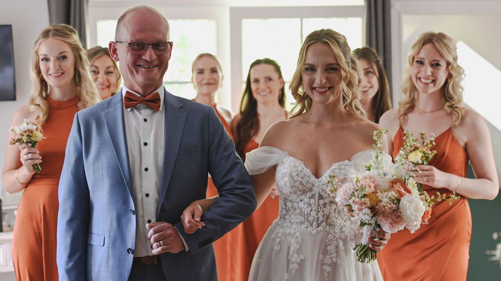 Wedding party poses in a row wearing orange bridesmaid dresses and formal attire at an indoor venue.
