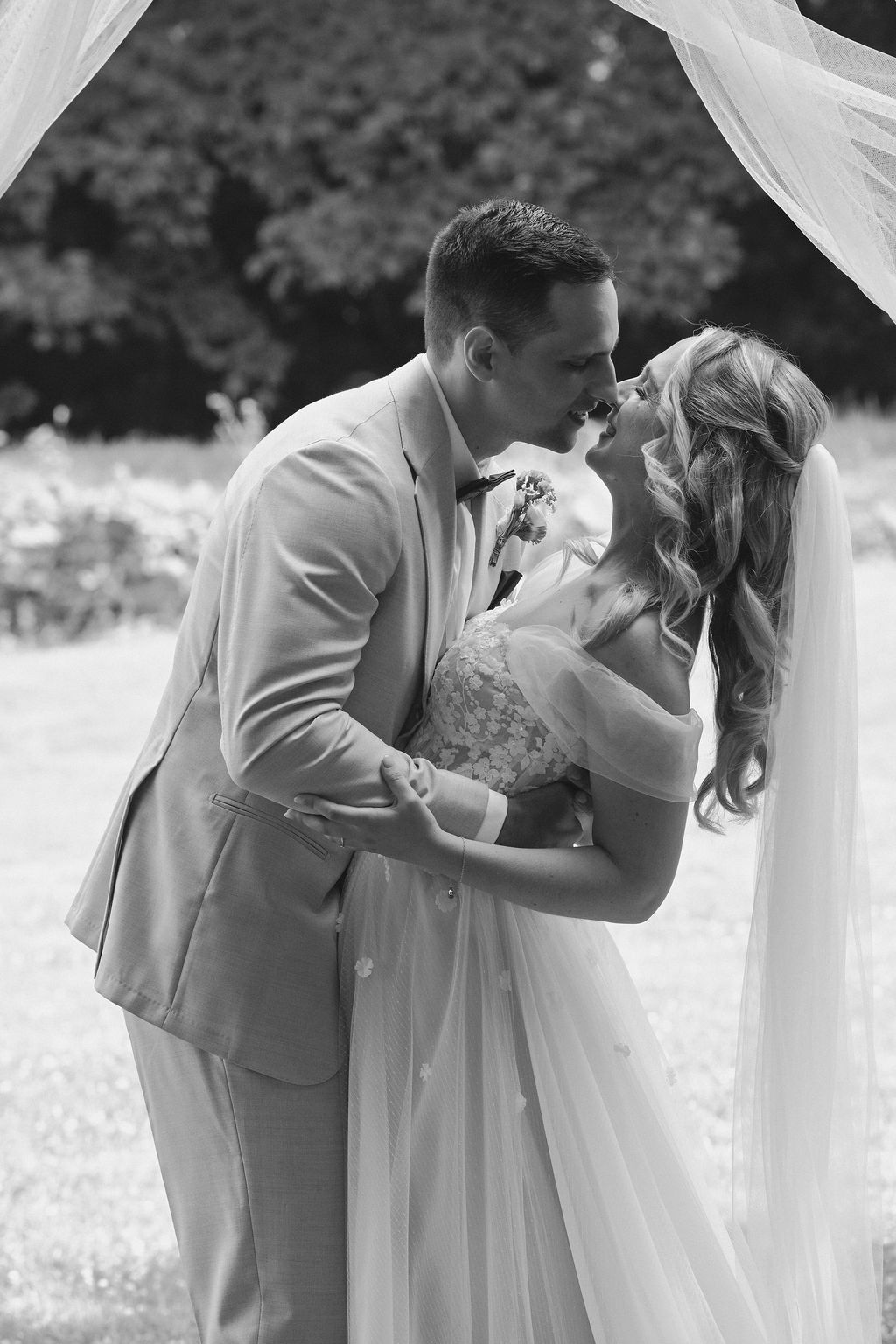 A romantic wedding couple shares a tender kiss under a white canopy outdoors in black and white photos.