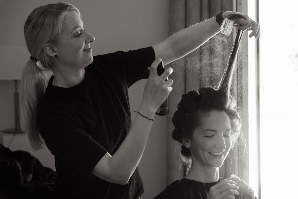 Black and white photo of a hairstylist styling a smiling woman's hair near a bright window.