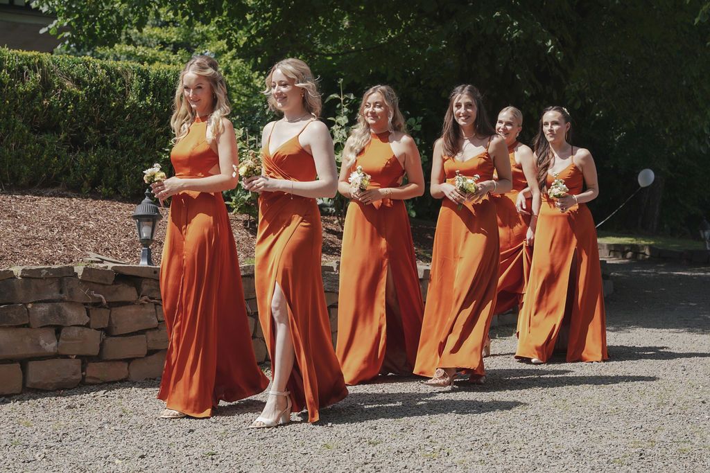 Bridesmaids in matching burnt orange dresses sit together on stone steps in an outdoor garden setting.