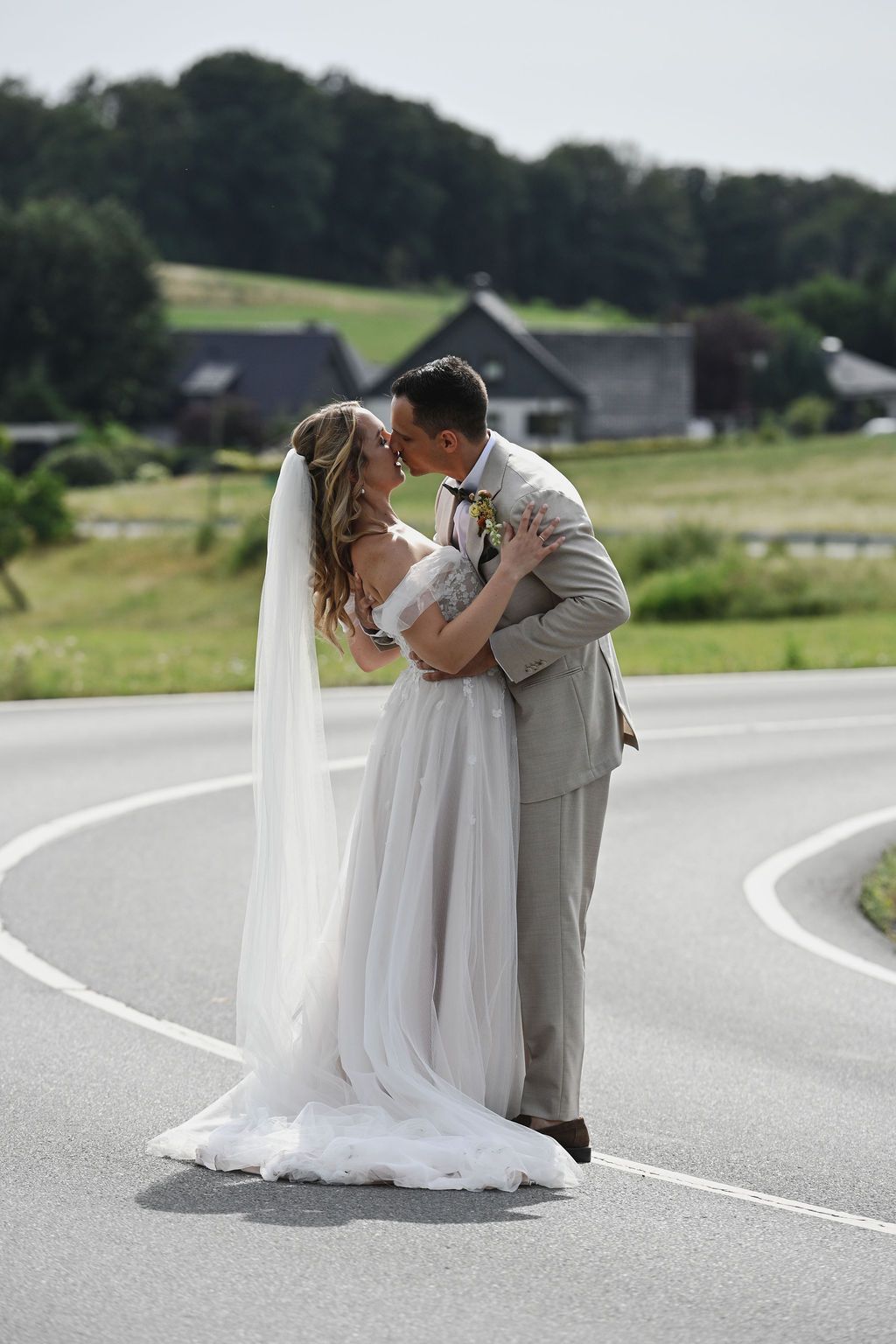 A couple in wedding attire share a romantic kiss on a winding country road.