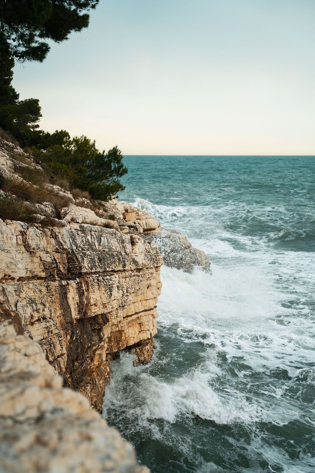 Dramatic limestone cliffs meet turquoise Mediterranean waters as waves crash against the rocky coastline during sunset.