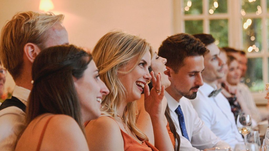 Wedding guests seated at reception tables smile and laugh during dinner celebration with string lights overhead.
