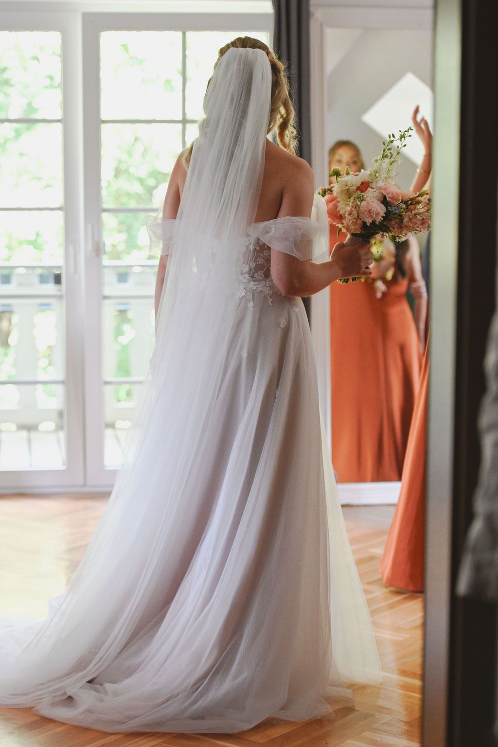 A bride in a flowing white wedding gown stands with bridesmaids wearing coral dresses in a sunlit room.