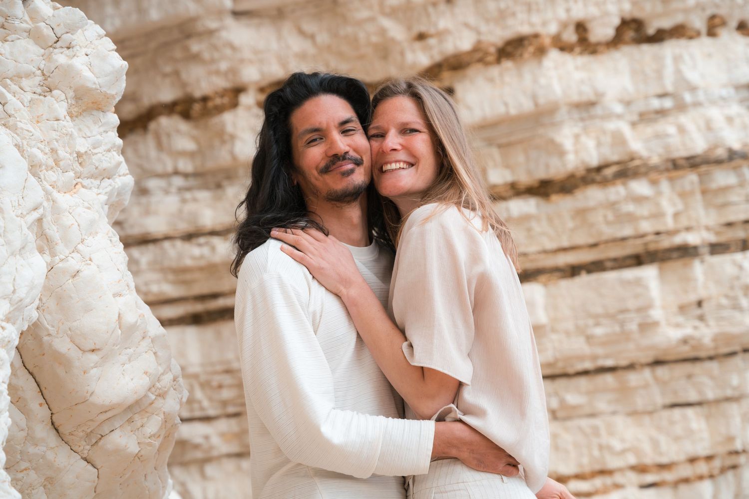 Happy couple embracing outdoors near sandy rock formations, wearing white outfits, smiling warmly in a romantic portrait.
