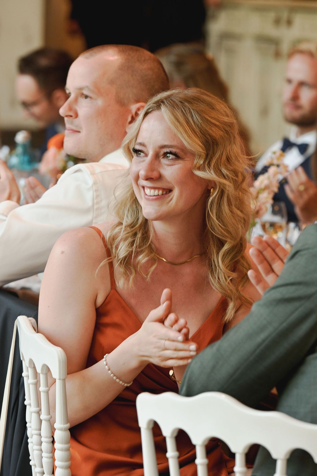 Wedding guest in orange dress laughs during reception while seated at table with other attendees.