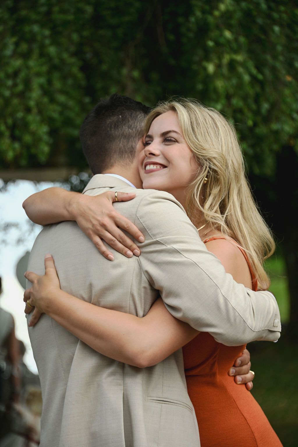 Two people embrace in a warm hug outdoors with greenery visible in the background.