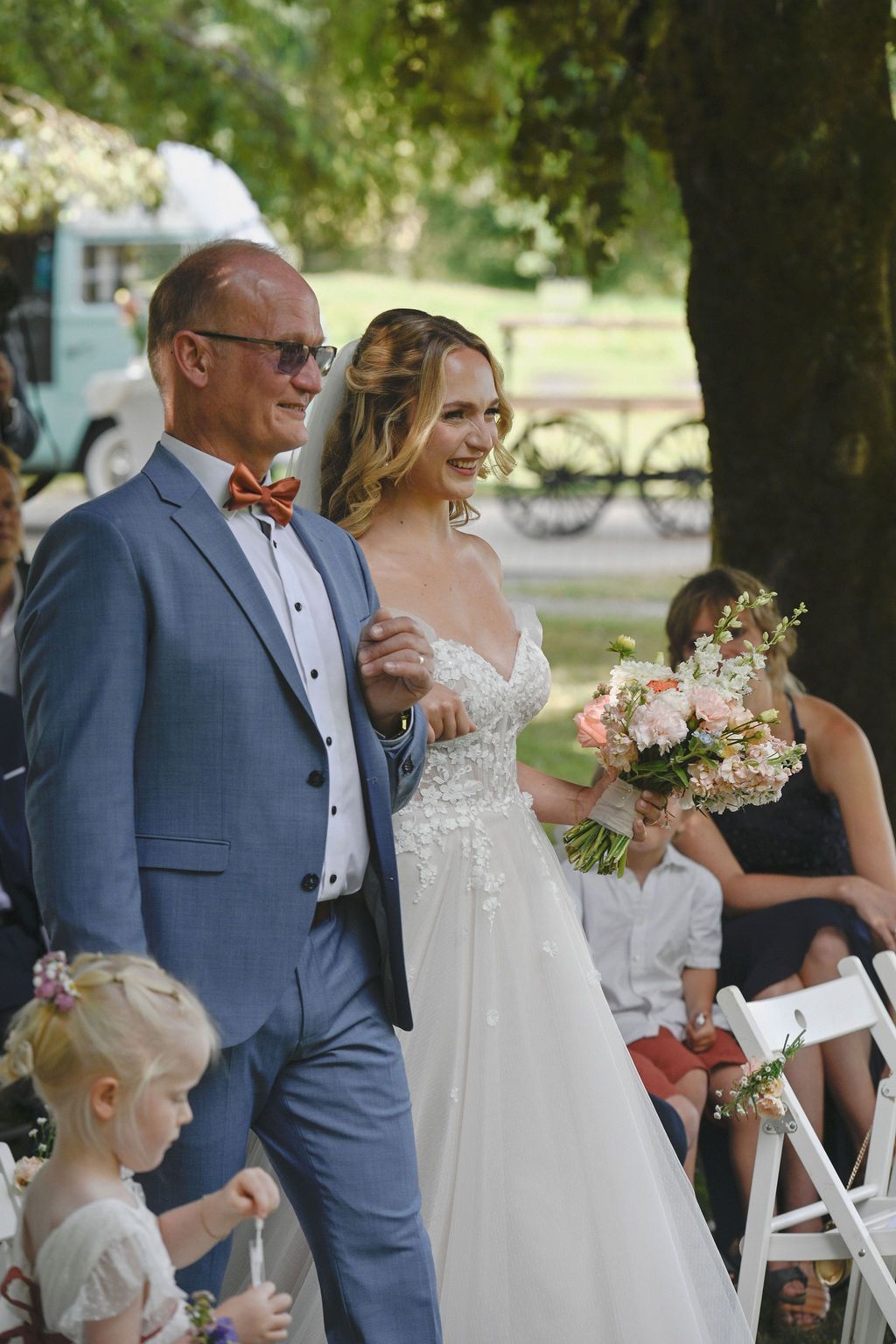 A wedding ceremony moment captured outdoors with the bride in a white dress and guests in formal attire.