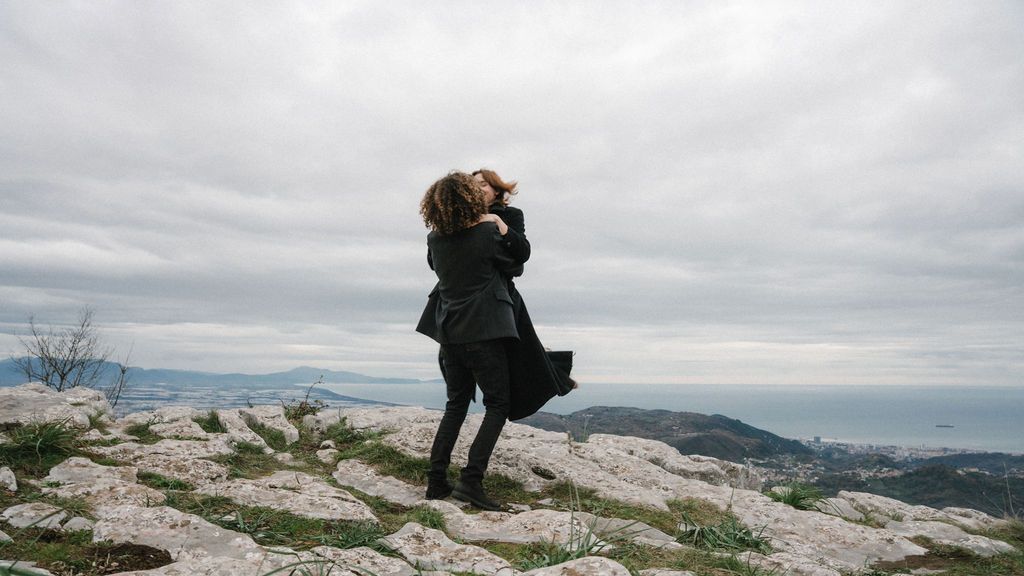 A couple embracing on a rocky mountain summit with cloudy skies and mountains in the background.
