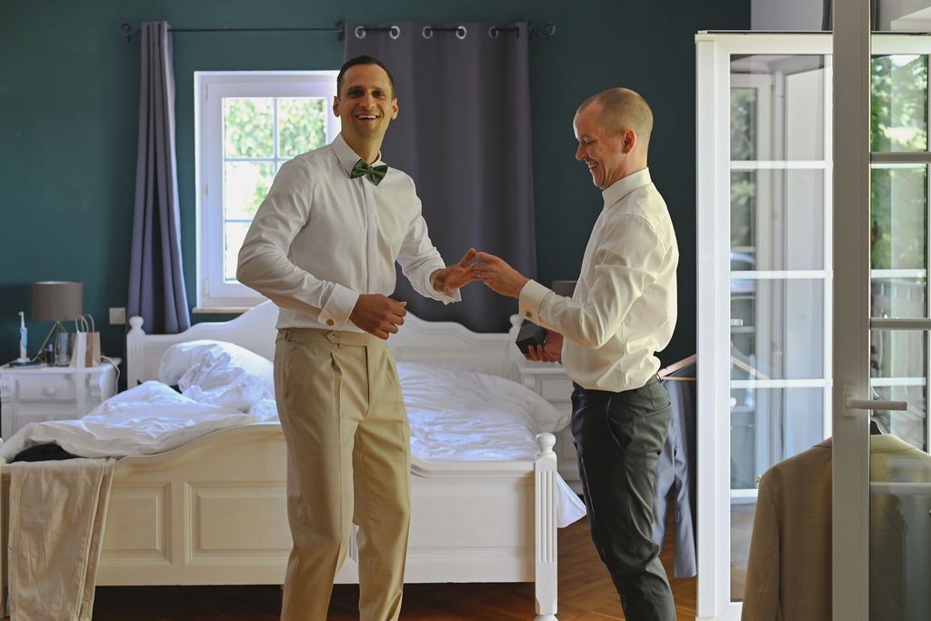 A group of people in formal attire stand and chat in an elegant white room with large windows.
